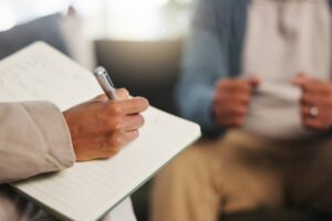 Book, therapy and hand of therapist writing notes for client with mental health evaluation