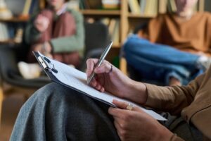 Young woman holding clipboard and writing notes during group therapy session with blurred multiethnic young participants