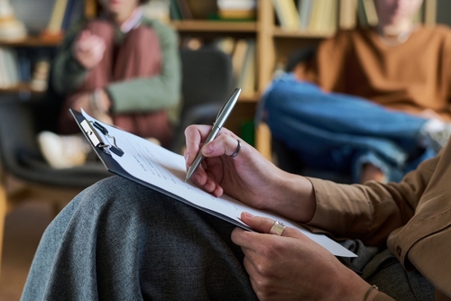 Young woman holding clipboard and writing notes during group therapy session with blurred multiethnic young participants