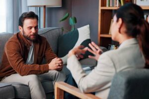 Psychologist and patient talk in therapy session. Clinician writes notes on a clipboard as a man talks about mental health during consultation. (2)