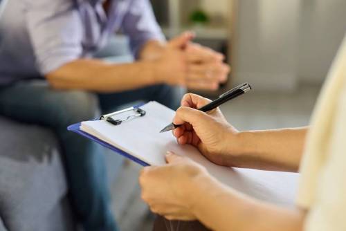 Psychologist and patient talk in therapy session. Clinician writes notes on a clipboard as a man talks about mental health during consultation.