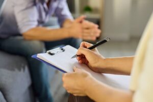 Psychologist and patient talk in therapy session. Clinician writes notes on a clipboard as a man talks about mental health during consultation.